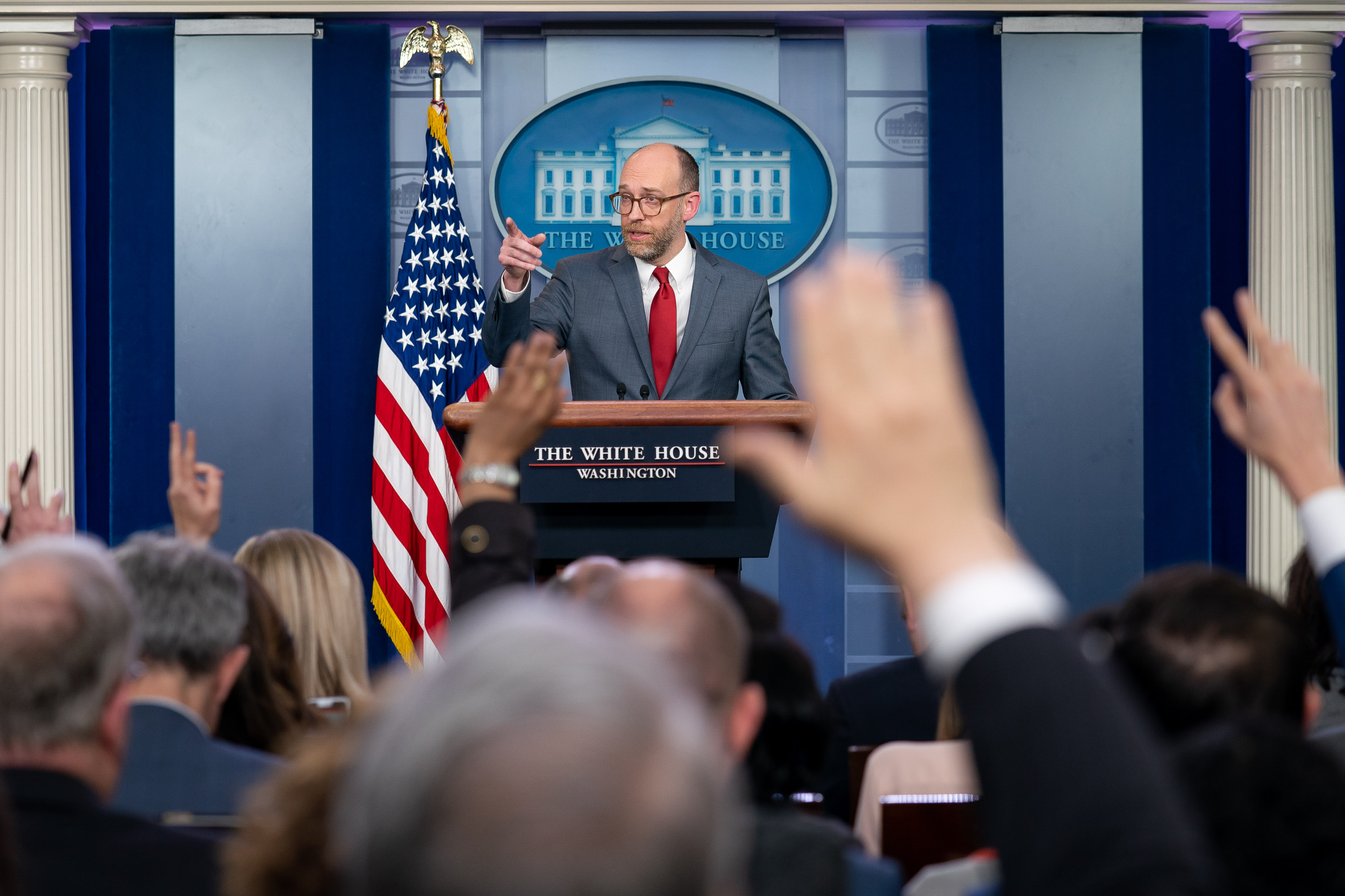Russell Vought talks with journalists during a March 2019 press briefing, back when he was acting director of the Office of Management and Budget. Standing behind a podium, in front of an American flag, Vought points to his right as if to call on one of the reporters, many of whom have raised their hands. Vought wears a dark blue-gray suit and red tie. He is white, balding and has brown-framed glasses.