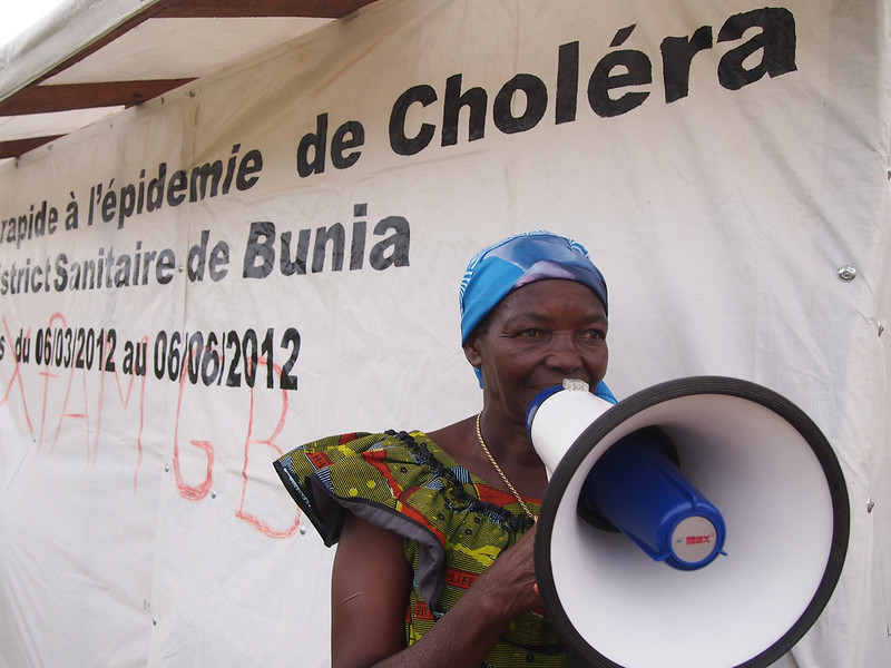 African woman with a bullhorn in front of a sign warning about cholera