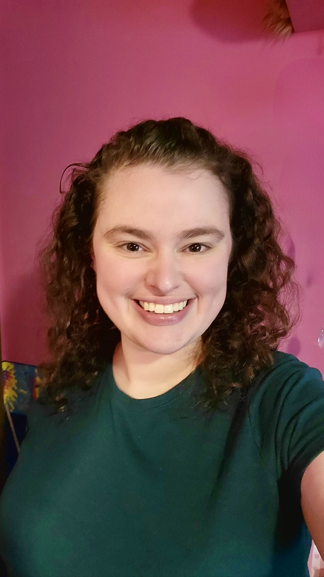 A woman with brown hair takes a selfie wearing a green shirt against a red background.