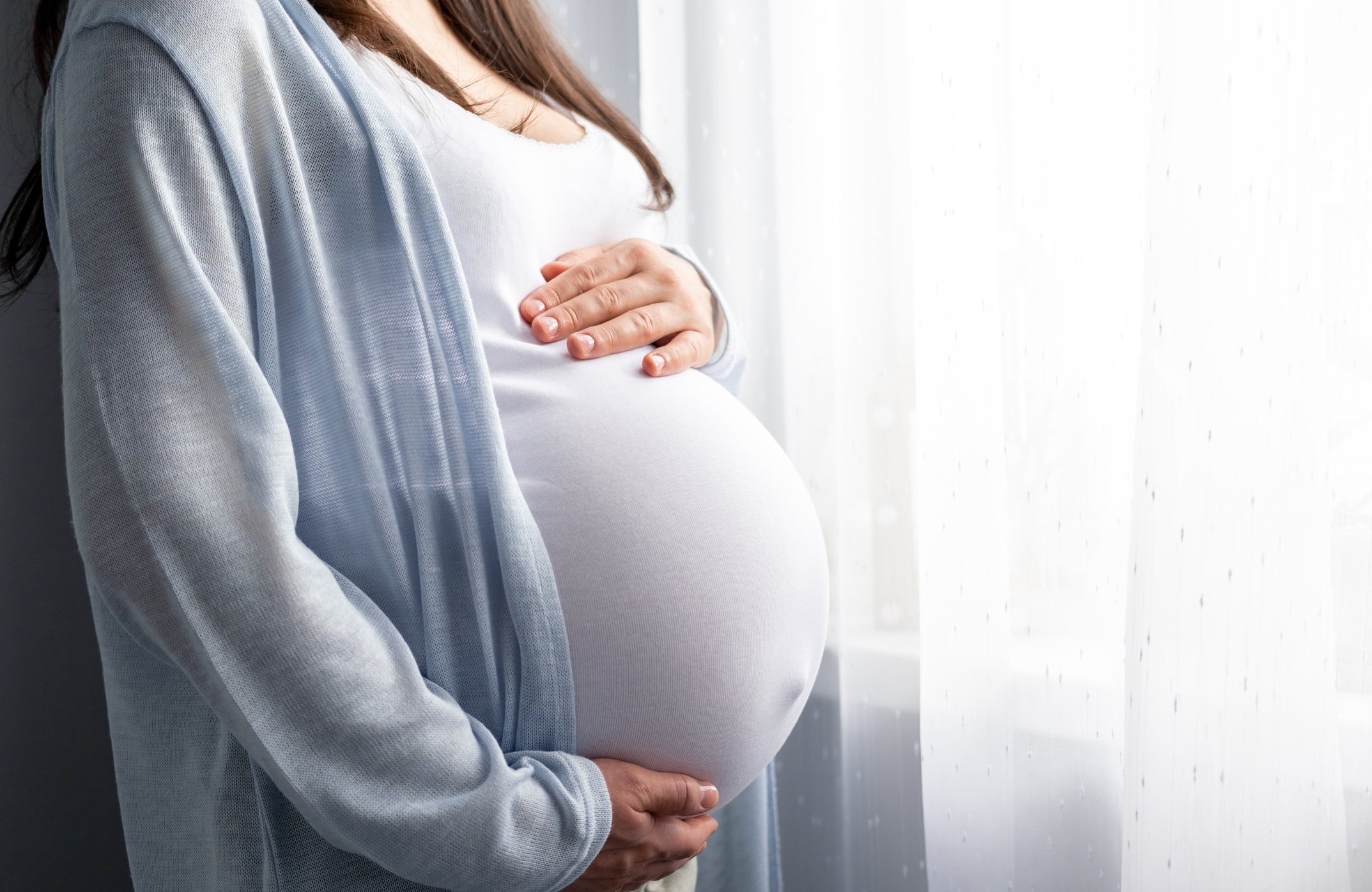Profile view of a pregnant woman in a white shirt and grey cardigan. Her face isn't visible. Left hand rests on top of her bump, and right hand cradles the bottom of the bump.
