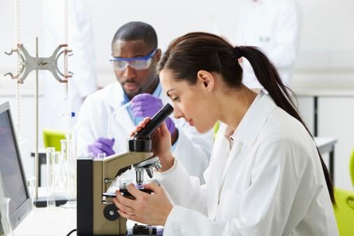 Two lab technicians, one holding a pipette and one looking into a microscope
