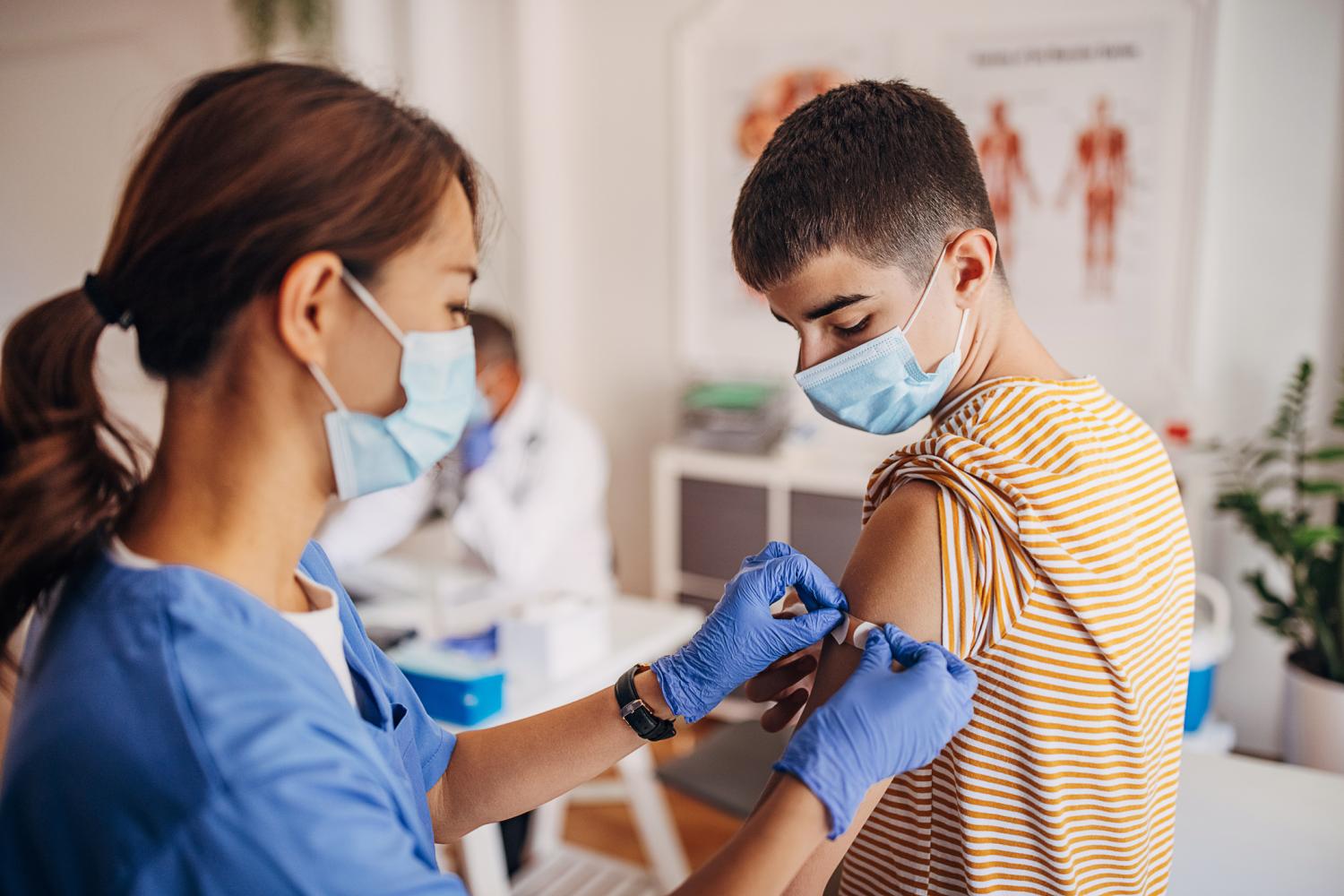 A school-age boy gets a vaccine in his arm from a health care provider in scrubs.
