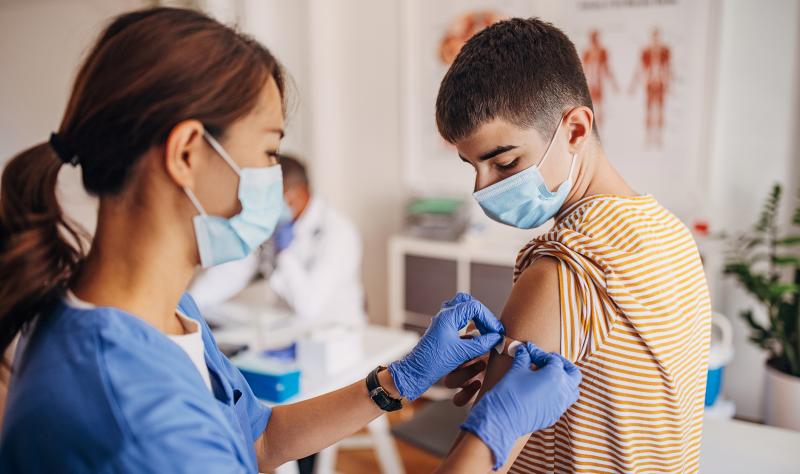 A school-age boy gets a vaccine in his arm from a health care provider in scrubs.