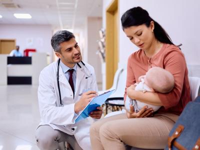 A woman in a pinkish-beige shirt holds a baby. She looks concerned. A smiling man in a white coat and tie kneels down to look at the baby. He holds a clipboard. They appear to be in a hospital.
