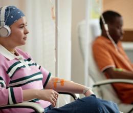Two seated adults, one of whom wears a bandana around their head, receive intravenous chemotherapy