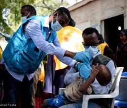 A healthcare worker administers medication to a small child being held by an adult in Somalia, while several other people look on.